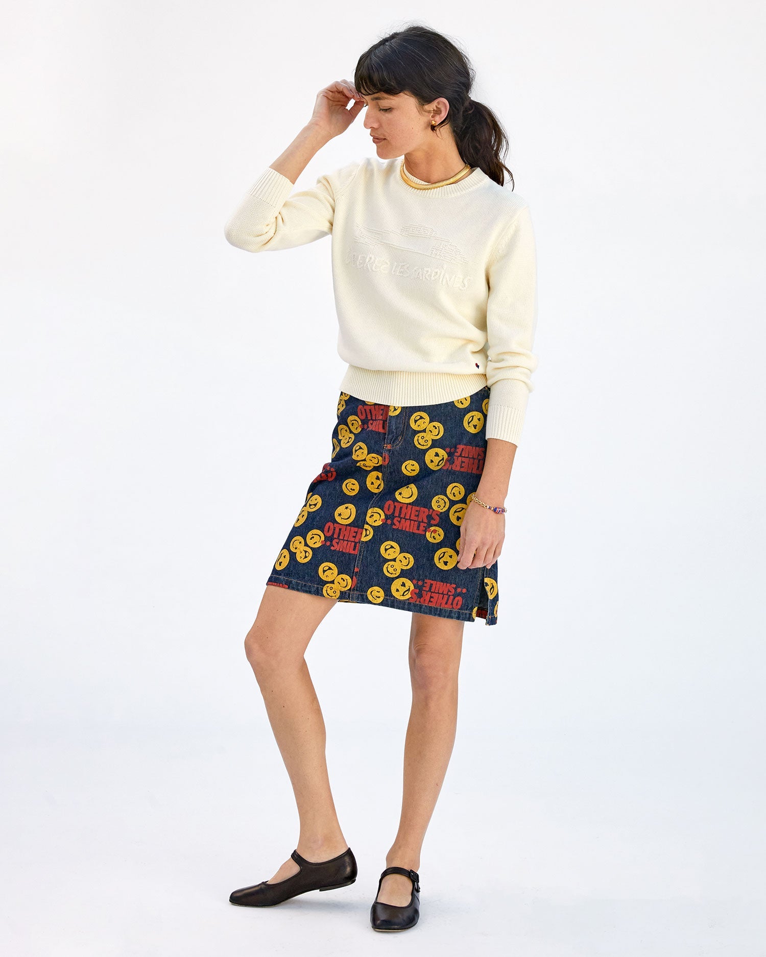danica wearing the cream liberez les sardines Classic Sweater with a skirt with emojis on it with black mary janes. she's brushing her hair behind her ear