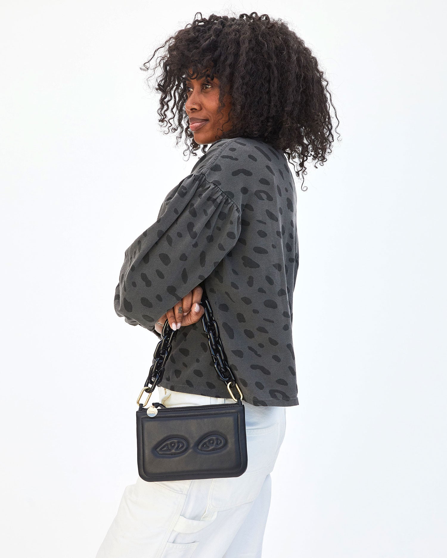 Mecca turned to the side wearing the Faded Black Jag Drop Shoulder Tee and holding  Black w/ Eyes Wallet Clutch w/ Tabs with the Black Resin Shortie Strap
