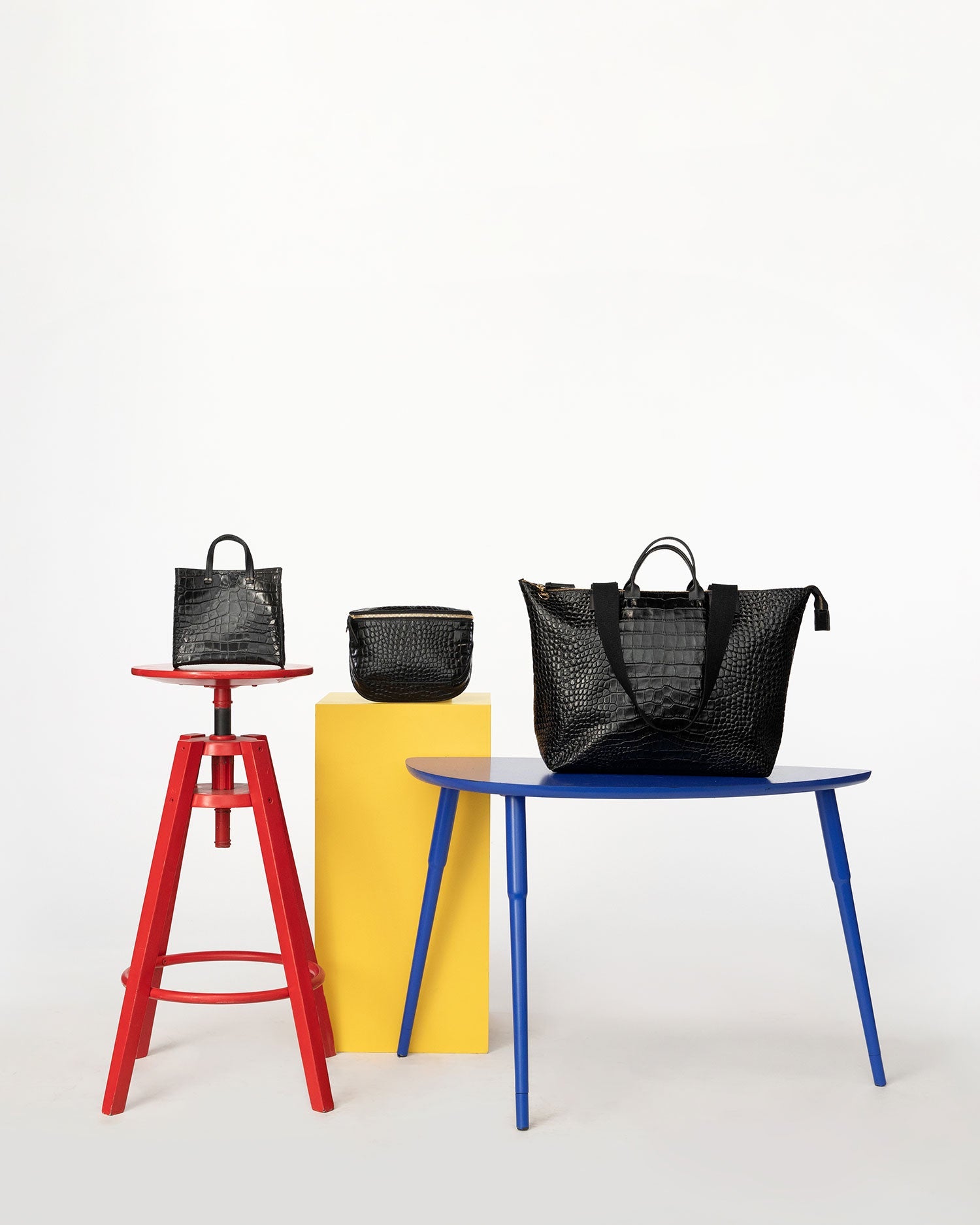 Black croco Simple Tote Bébé on a red stool, the black croco fanny pack on a yellow stool and the black croco le zip sac on a blue table 