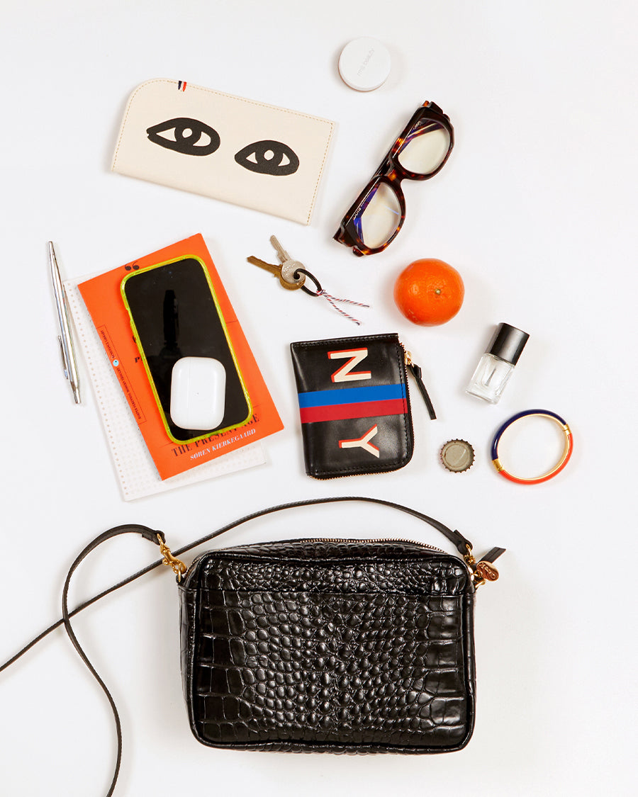 Black Croco Marisol w/ front pocket pictured with the items that can fit inside of it. These items include, a glasses case, a pair of glasses, a clementine, a set of keys, a corner zip wallet, a bracelet, a nail polish, a small notepad, a small book, a pen, an iphone and a pair of airpods