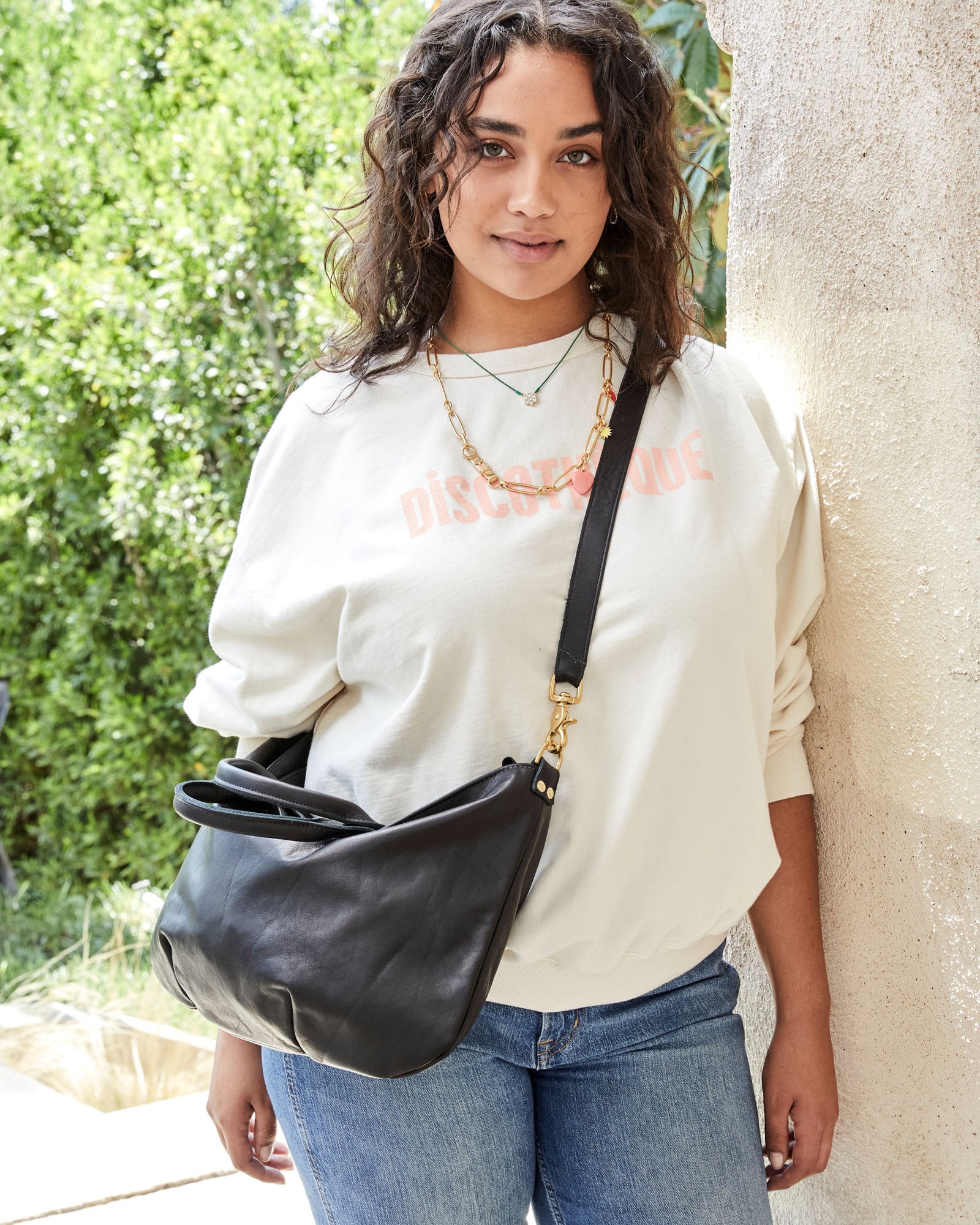 Claudia Wearing the Black Lamba Messenger Crossbody, Leaning Against a Wall